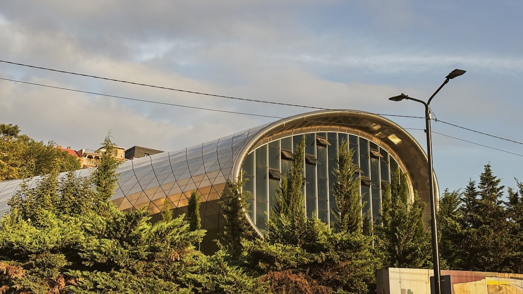 The sleek tubular design of Tbilisi’s modern concert hall catches the warm evening light, its glass and steel facade standing out against the surrounding greenery and skyline.